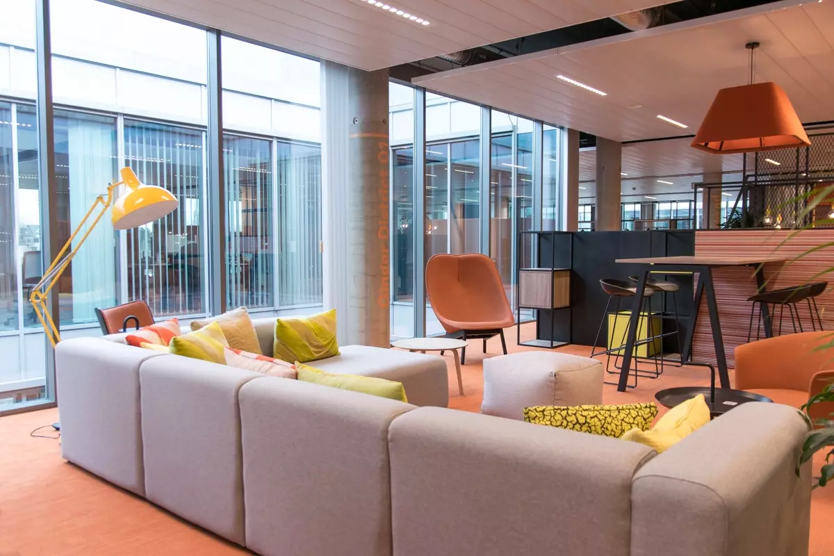 An open-plan office environment with a comfortable seating area, featuring a grey sofa, colourful cushions, and orange seats.