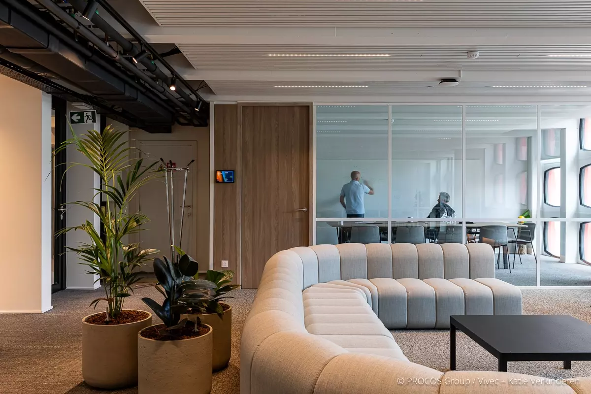 Modern office with curved sofa, plants and glass meeting room. Two people work together at a whiteboard.